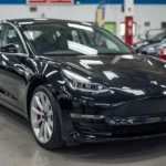 Tesla used car prices dropping in the US market showing a Tesla Model 3 parked at a dealership with price tags highlighting lower resale values.