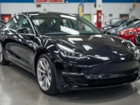 Tesla used car prices dropping in the US market showing a Tesla Model 3 parked at a dealership with price tags highlighting lower resale values.