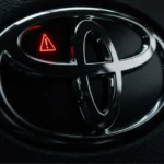 A sleek, modern Toyota steering wheel emblem with the reflection of a red dashboard warning light, symbolizing the urgent Toyota Recall 2025 for a critical airbag sensor defect.